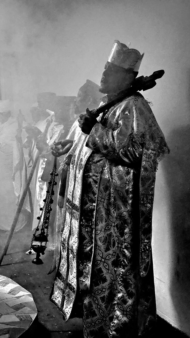 A black-and-white photograph of a vested clergyman in an ornate crown and brocade robes, swinging a censer amid rising incense smoke during a procession.