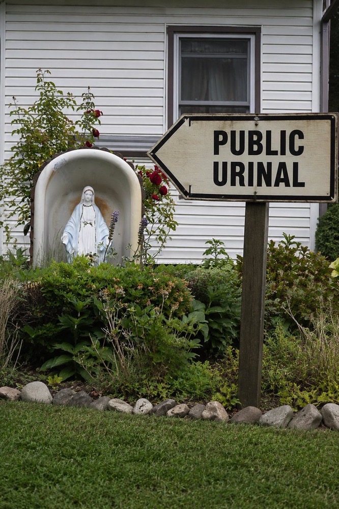 A statue of the Theotokos in a garden shrine, with a sign reading PUBLIC URINAL pointing directly at her.